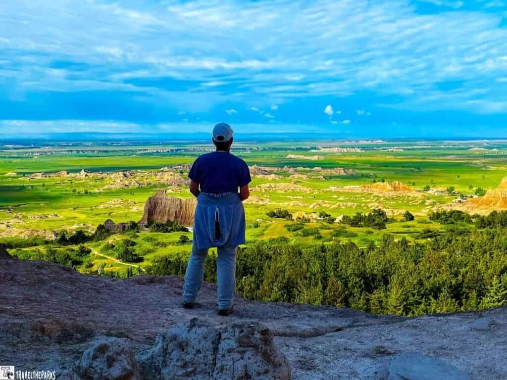Ultimate South Dakota and North Dakota Road Trip: 15 Unforgettable Stops: A person stands on a rocky ledge, overlooking a vast green landscape with hills and blue skies.