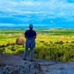 Ultimate South Dakota and North Dakota Road Trip: 15 Unforgettable Stops: A person stands on a rocky ledge, overlooking a vast green landscape with hills and blue skies.