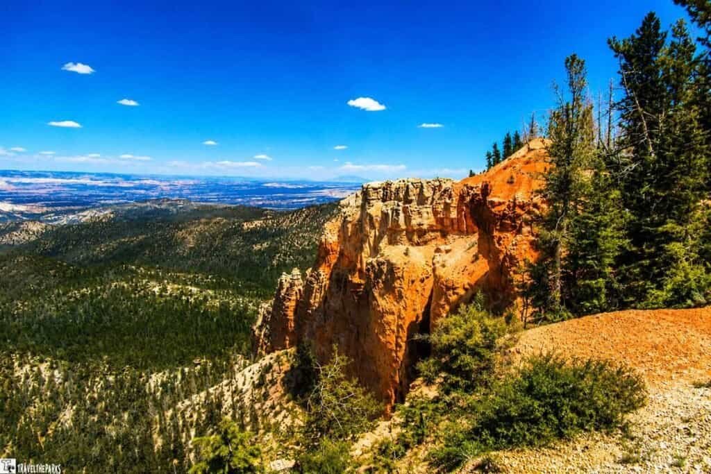 A panoramic view of Bryce Canyon featuring a rugged orange cliff in the foreground, surrounded by green trees, under a blue sky with scattered clouds.