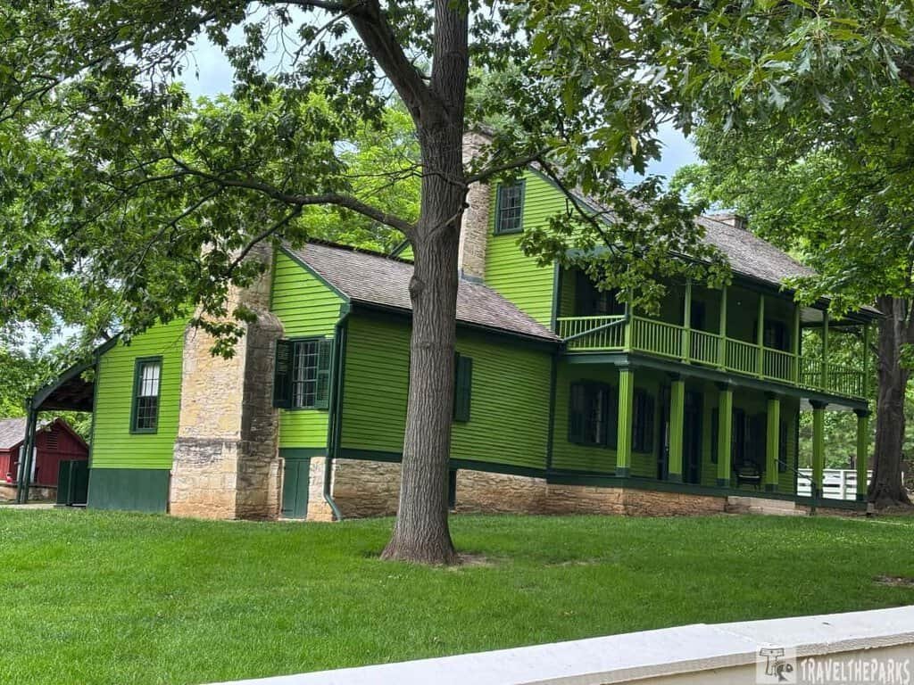 Historic two-story green building with stone lower half, surrounded by trees and grass; a red barn is visible in the background.