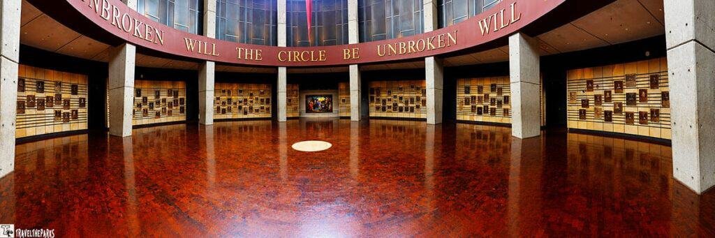 Interior view of a circular room with wooden panels displaying plaques and the text "WILL THE CIRCLE BE UNBROKEN" above. A polished floor reflects natural light.


