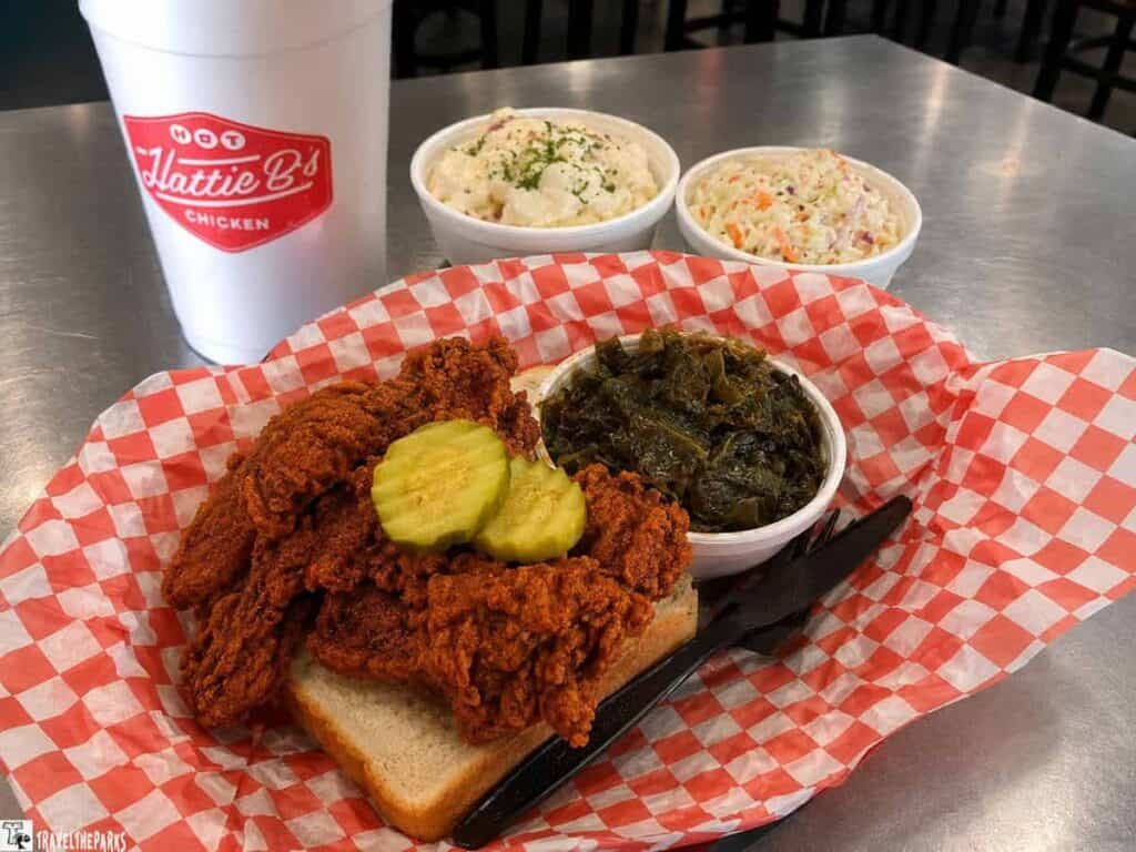 A plate of spicy fried chicken with pickles on white bread, accompanied by collard greens and two creamy salads; a cup from Hattie B's Chicken is in the background.

