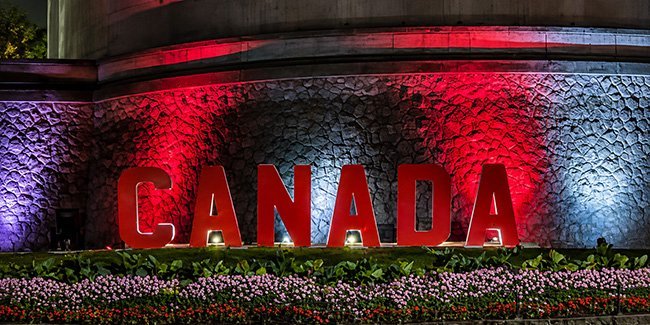 Large red “CANADA” letters stand before a textured stone wall lit in red, purple, and blue, with colorful flower beds below.