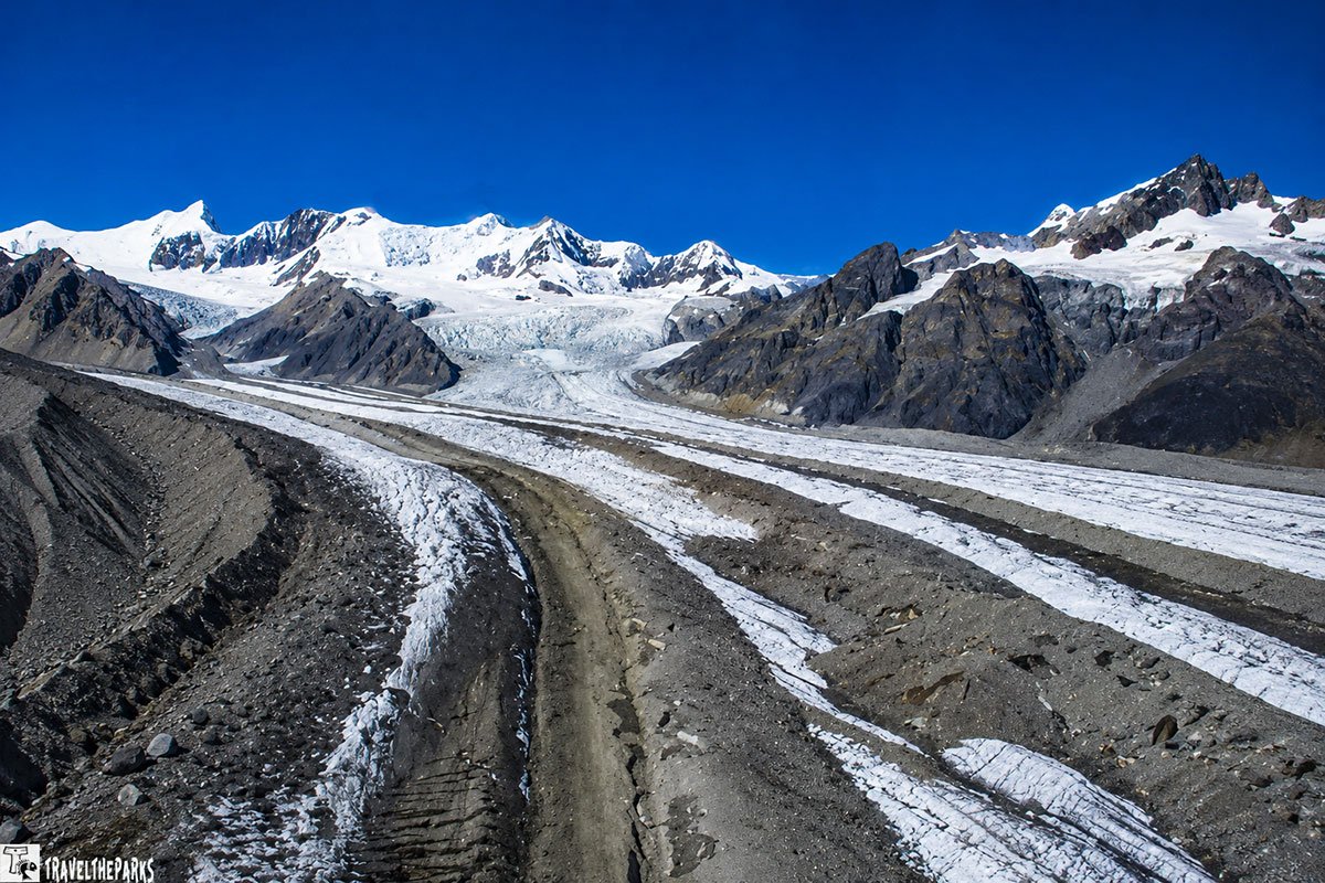 A winding dirt path leads through rocky terrain towards Kennicott Glacier and Root Glacier, framed by snow-capped mountains under a clear blue sky.