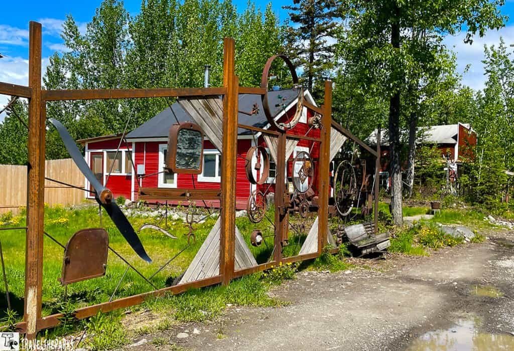 A decorative metal gate featuring an airplane propeller and gears, with a red wooden building and green landscape in the background.