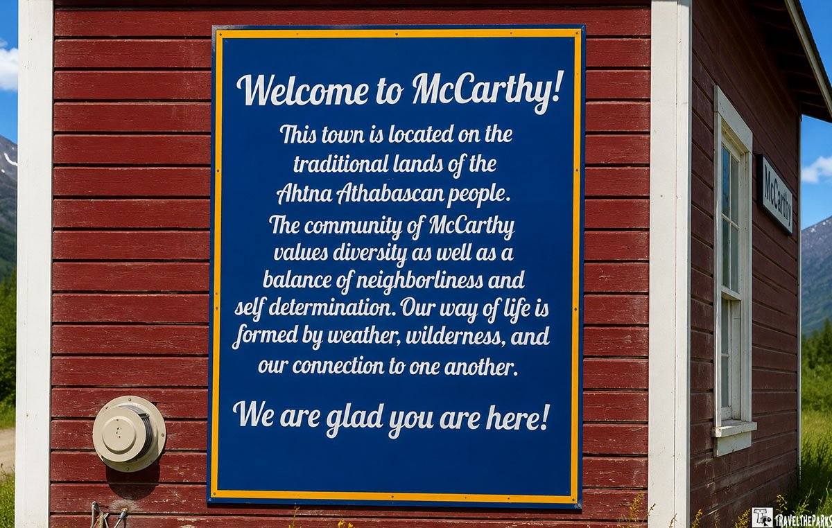 A welcome sign in blue with white text and a yellow border, mounted on a red wooden wall, stating the town's cultural significance and values.
Transcribed Text:
Welcome to McCarthy!
This town is located on the traditional lands of the Ahtna Athabascan people. The community of McCarthy values diversity as well as a balance of neighborliness and self-determination. Our way of life is formed by weather, wilderness, and our connection to one another.
We are glad you are here!