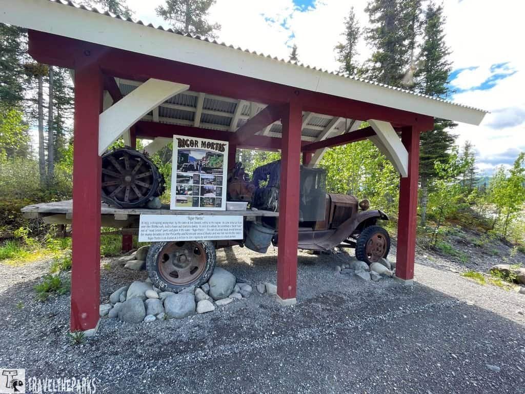 An old vehicle displayed under a rustic shelter, with an informational poster about "Rigor Mortis" in front, set against a backdrop of greenery.
