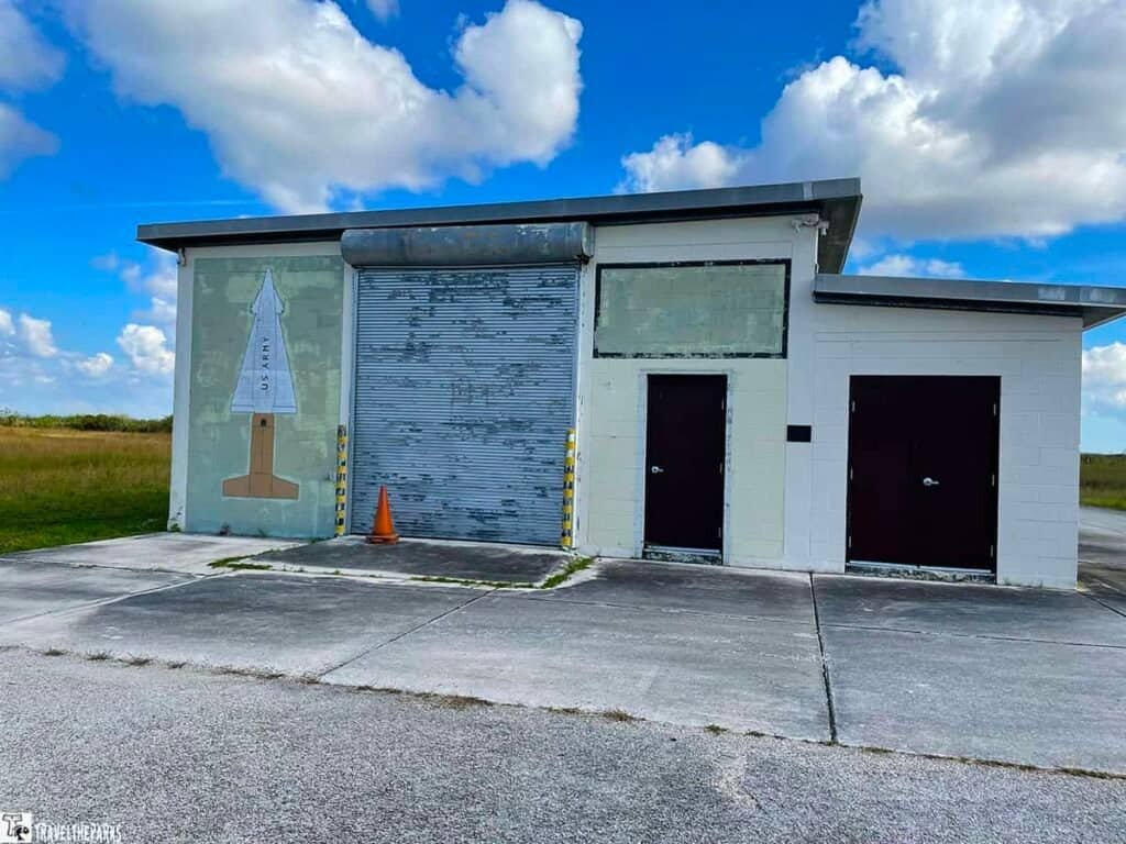 Nike Missile Base Building-A small building with a flat roof and a central corrugated metal door, featuring a faded mural of a rocket. An orange cone stands in front.