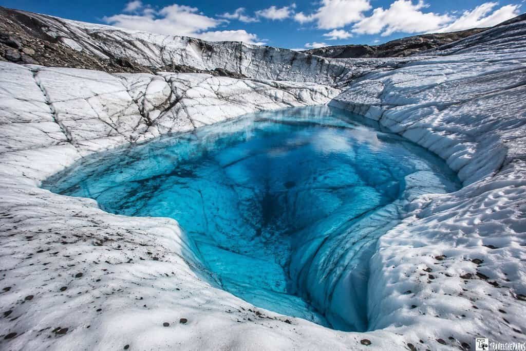 Bright blue glacial pool surrounded by textured white ice, with scattered dark rocks and a clear sky above.