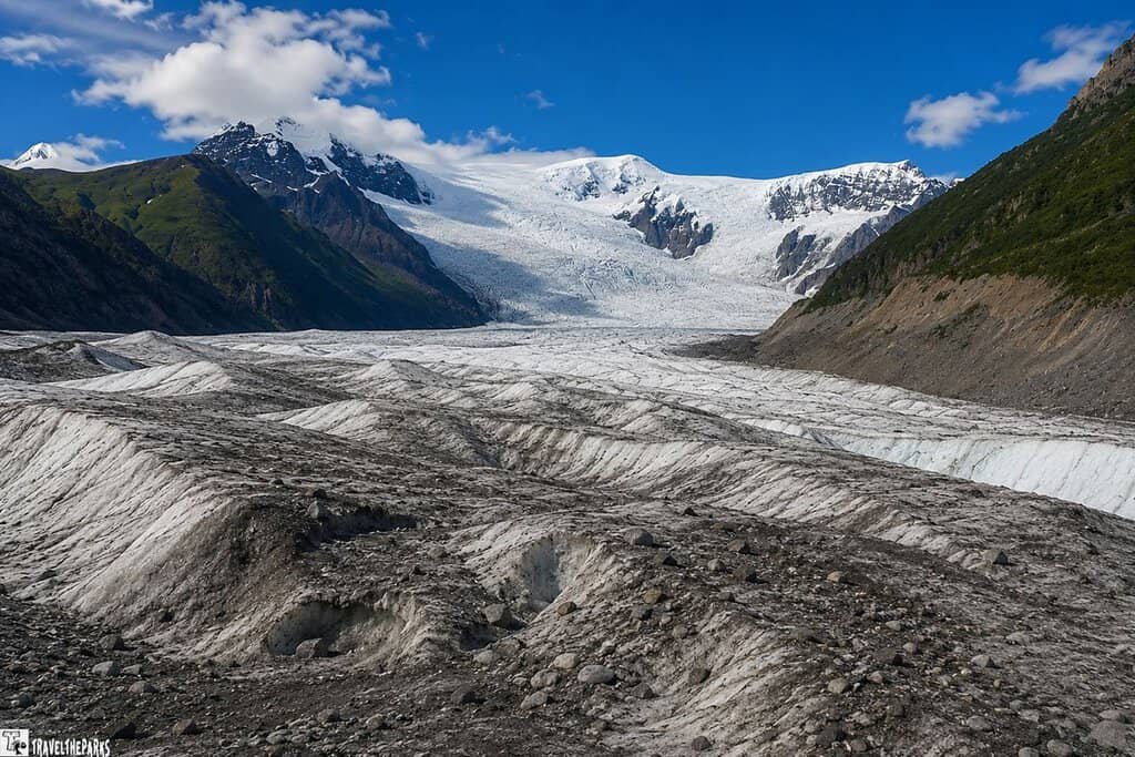 A view of the Kennicott and Root Glaciers in Alaska, featuring white glacial ice and rocky terrain, with rugged mountains and a blue sky above.
