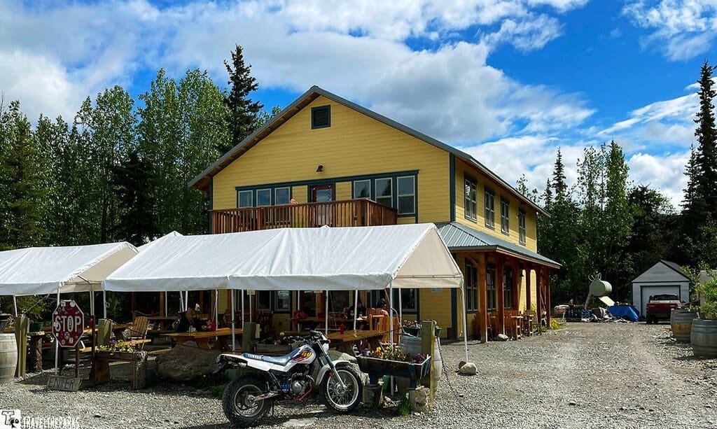 The Potato Restaurant McCarthy Alaska: A yellow two-story building with a metal roof, canopies, and a motorcycle in front, surrounded by trees.