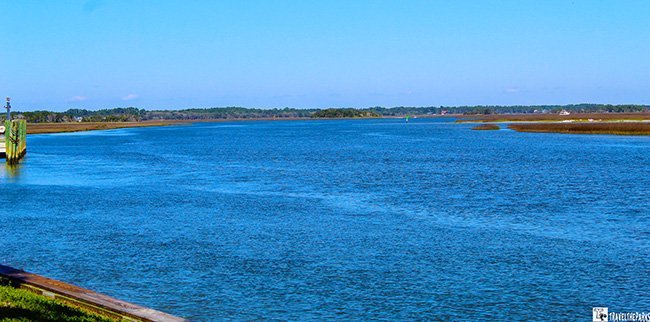 Wide view of a blue waterway bordered by low marshland under a clear sky.
