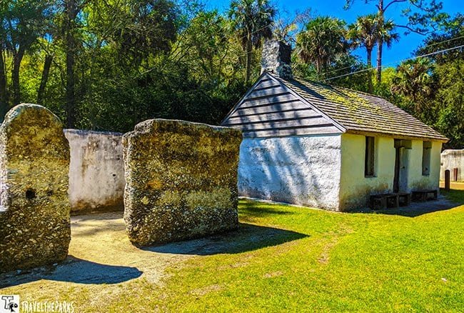 a historical site at Kingsley Plantation. In the foreground, there are remnants of tabby slave quarters, characterized by rough-textured walls made from a mix of lime, sand, and oyster shells. These structures are weathered and predominantly gray. Behind these ruins stands a small, whitewashed building with a steeply pitched wooden roof. The building has a chimney protruding from its roof and two small, rectangular windows. The surrounding area is a grassy lawn with patches of light brown soil. Tall trees with lush green foliage are visible in the background under a clear blue sky.